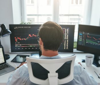 Trader acompanha gráficos do mercado financeiro em plataforma de negociação, representando o modelo de mesa proprietária e a formação profissional no mercado de trades.