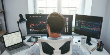 Trader acompanha gráficos do mercado financeiro em plataforma de negociação, representando o modelo de mesa proprietária e a formação profissional no mercado de trades.