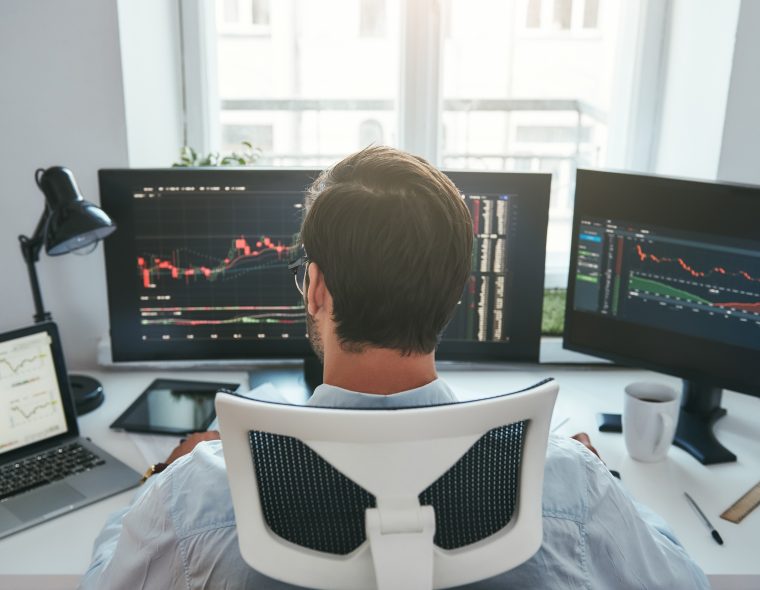 Trader acompanha gráficos do mercado financeiro em plataforma de negociação, representando o modelo de mesa proprietária e a formação profissional no mercado de trades.